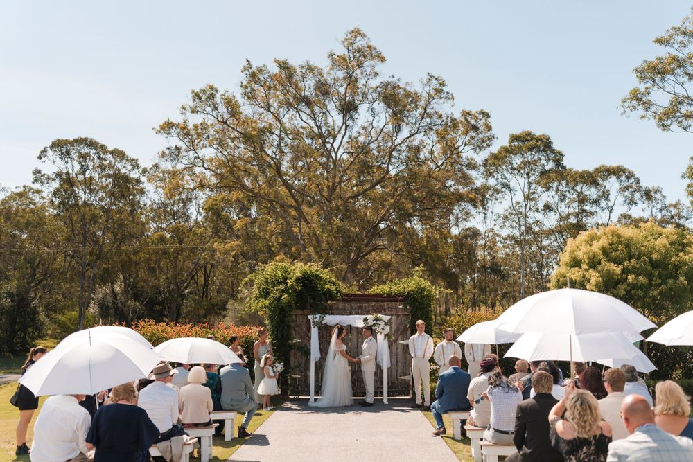 Outdoor wedding ceremony in a garden setting with guests holding white umbrellas under eucalyptus trees.