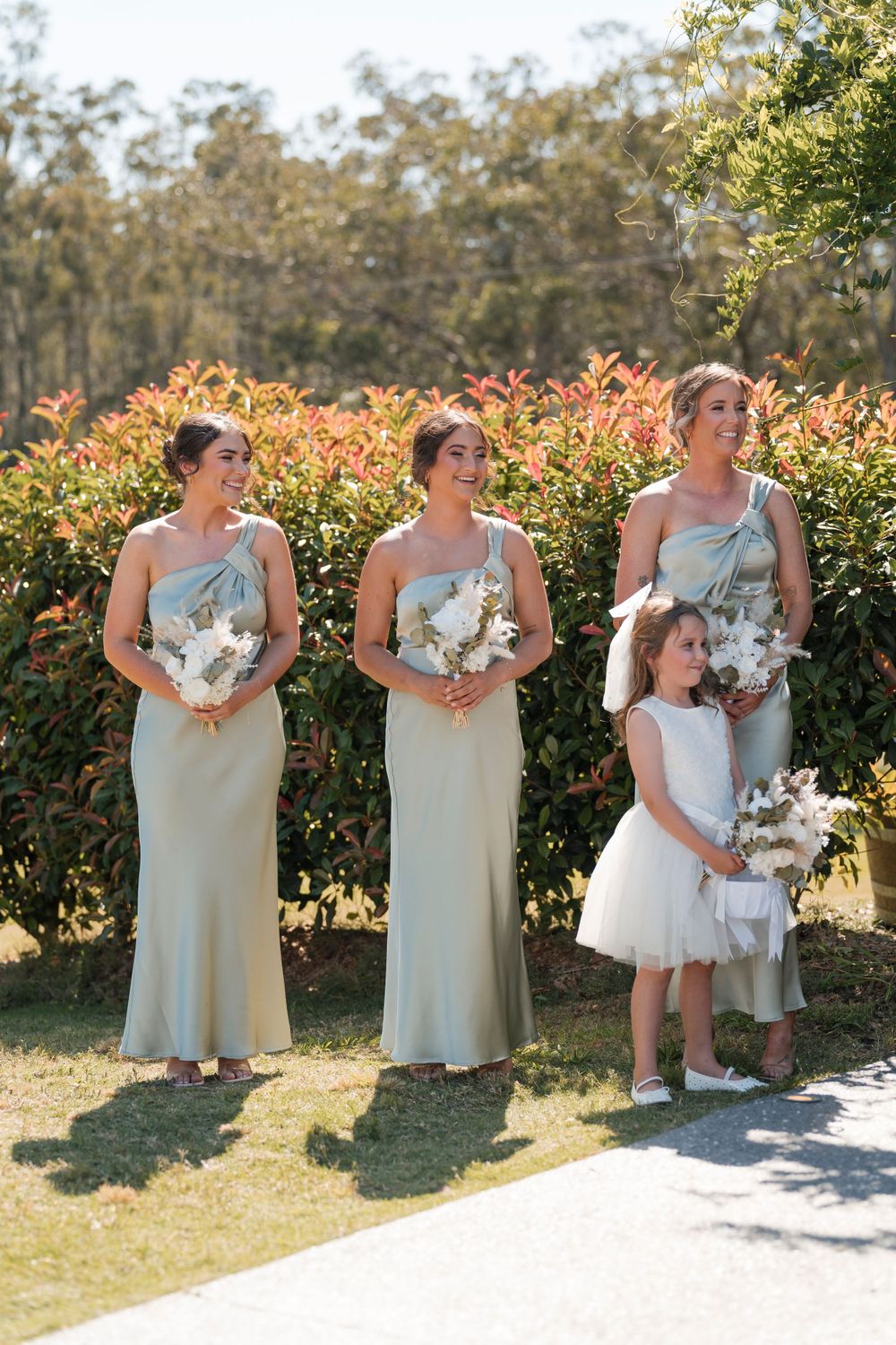 Wedding party members in sage green dresses hold white bouquets while standing in front of blooming shrubs on a sunny day.