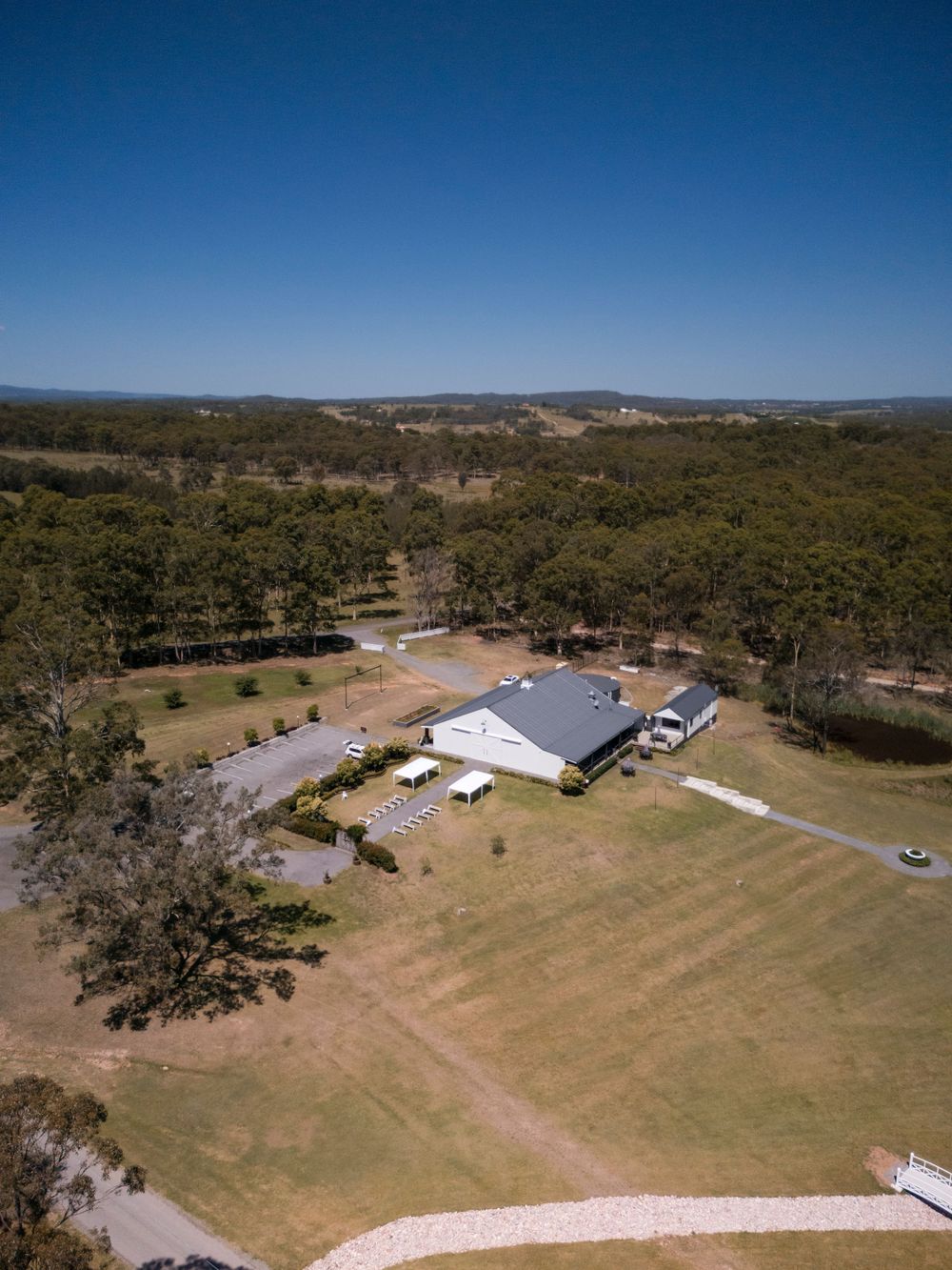 Aerial view of a white barn building surrounded by grassy fields and forest in a rural countryside setting.