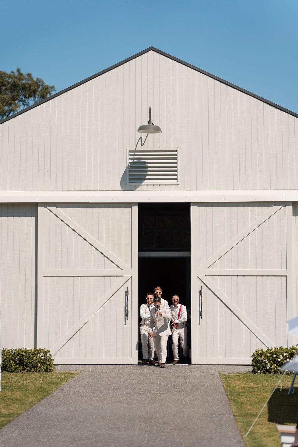 A white barn-style wedding venue with large sliding doors frames three people standing in formal attire.
