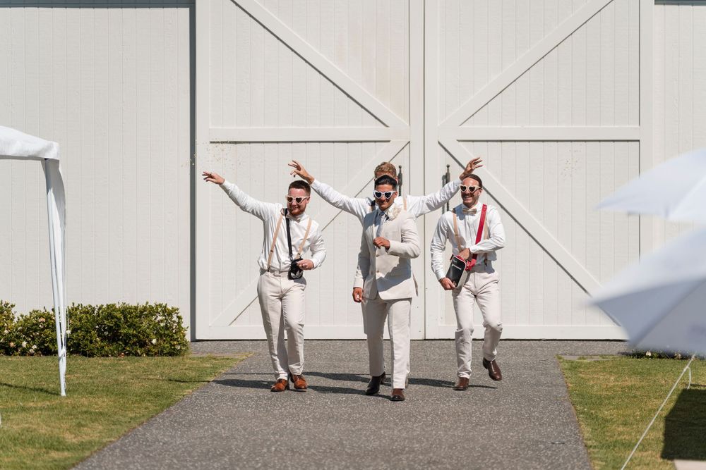 Three wedding guests in light-colored attire dance and celebrate together on a pathway outside a white barn.