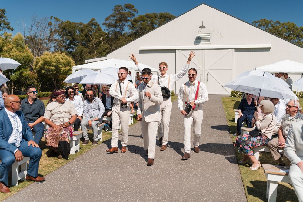 A wedding party in cream-colored attire walks down an outdoor aisle with guests seated on either side and a white barn behind them.