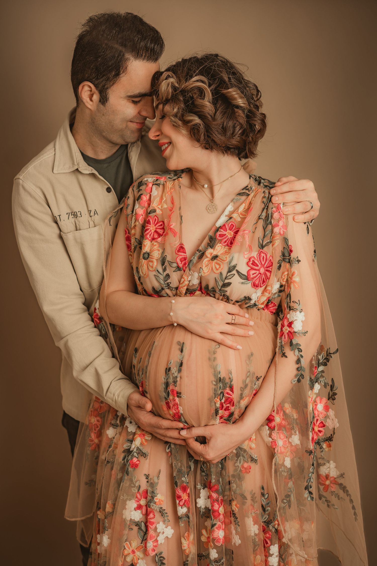 Maternity portrait featuring a floral dress and tender embrace in warm studio lighting.