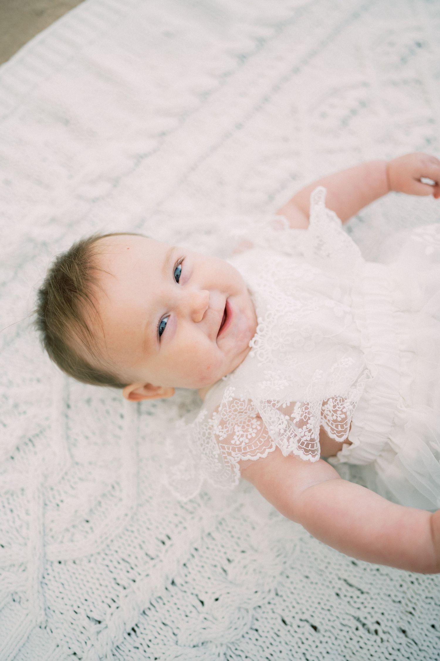 Baby wearing delicate white lace dress lies on soft blanket with joyful expression.
