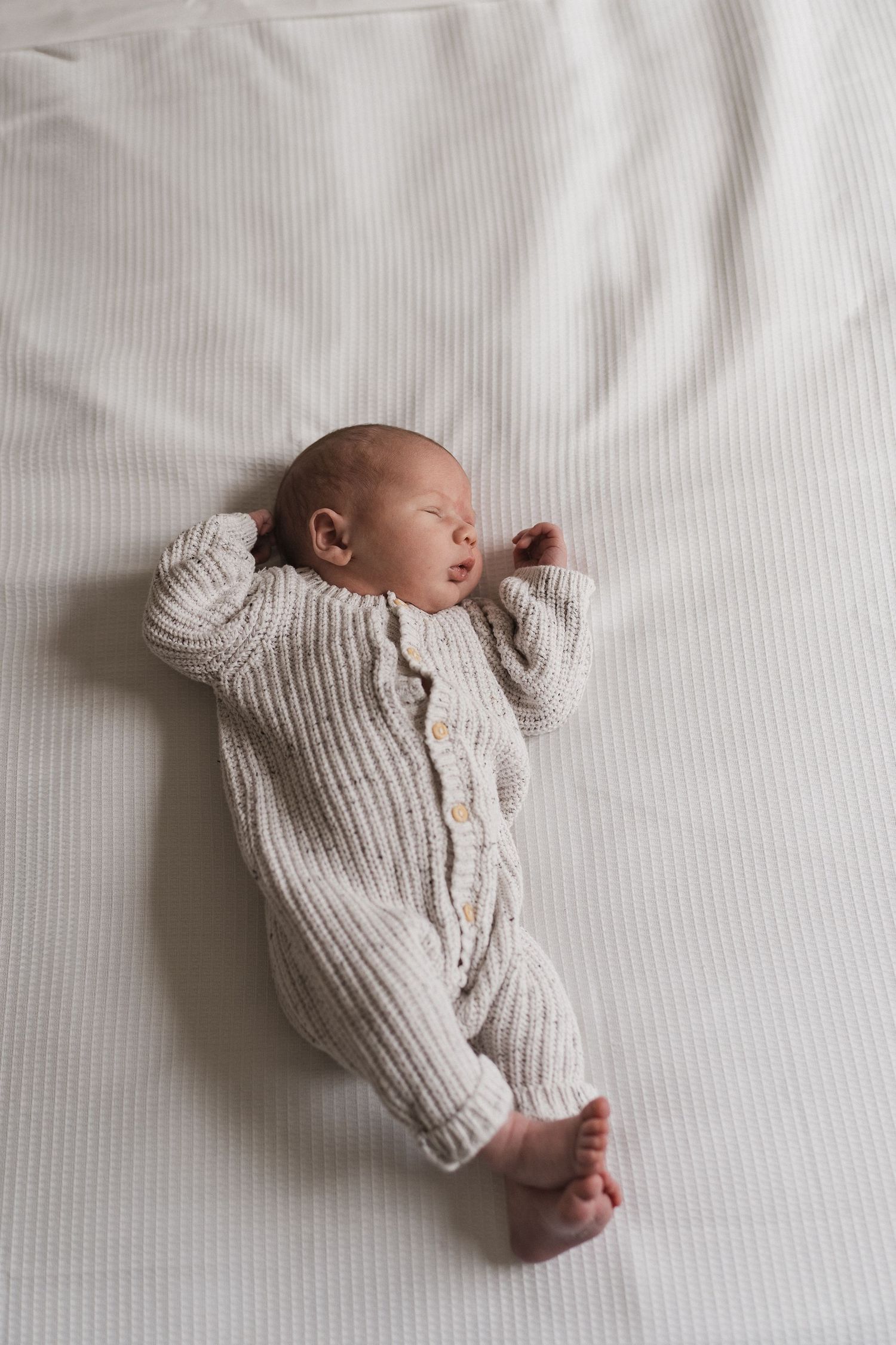 Sleeping newborn in cozy white knitted onesie lying on white textured surface.