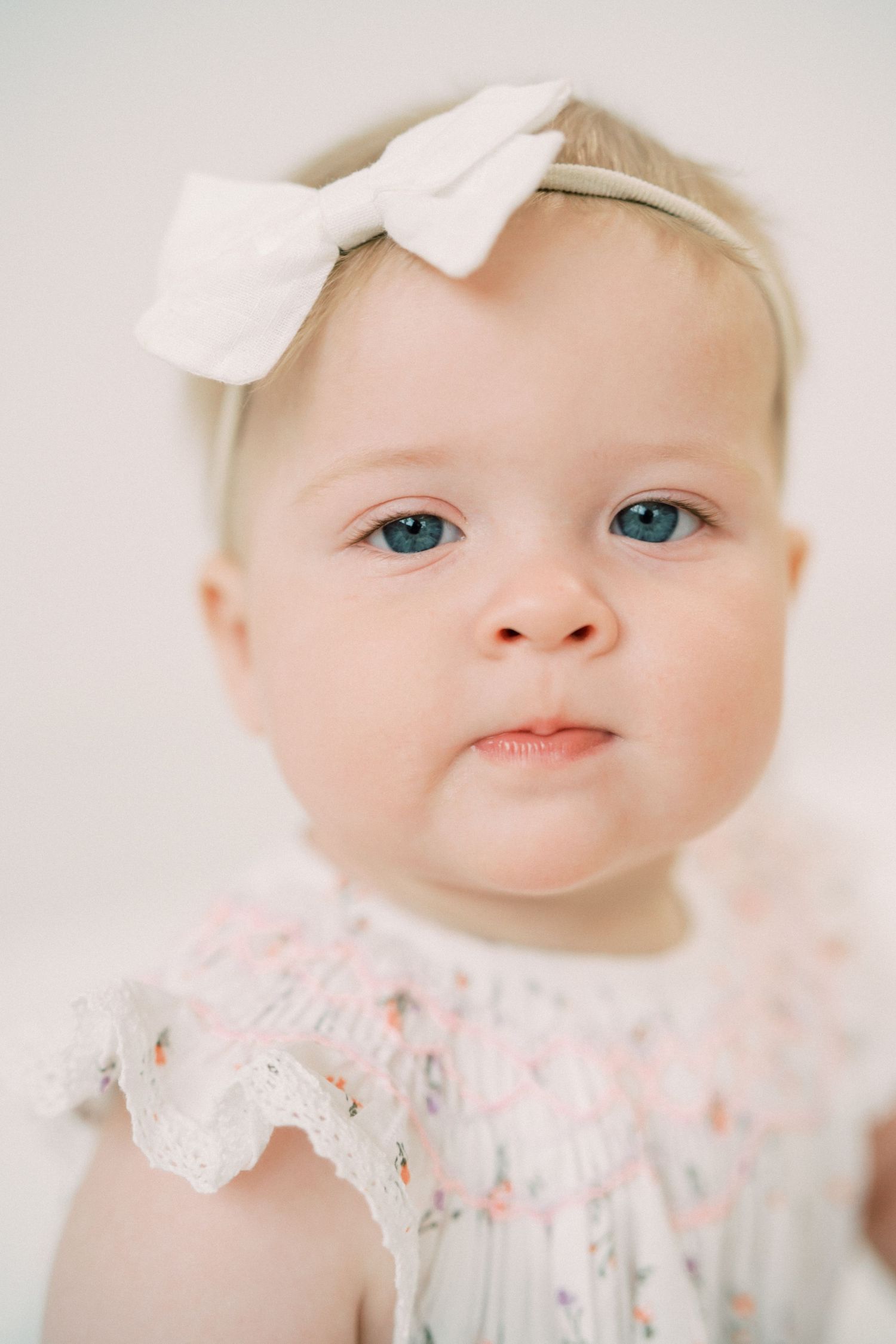 Baby wearing floral smocked dress and white bow headband looks at camera with bright blue eyes.