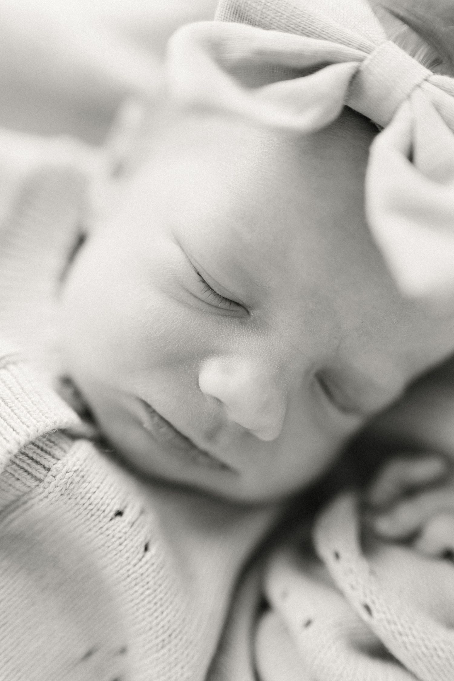 A peaceful black and white close-up photograph of a sleeping baby wearing a light bow headband.