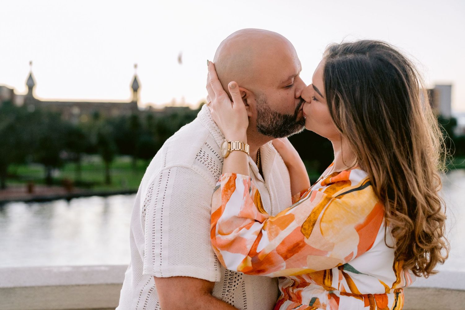 A romantic couple shares a kiss at sunset by a waterfront with urban buildings in Downtown Tampa.