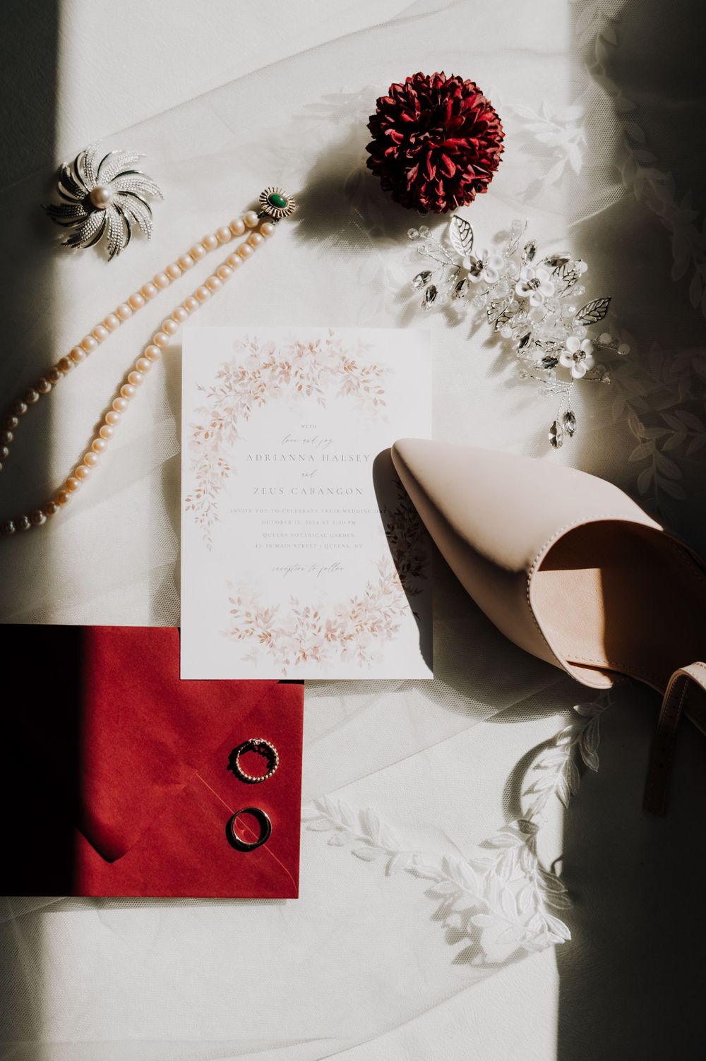Wedding details flatlay with rings, invitation, pearls and red accents on white background.