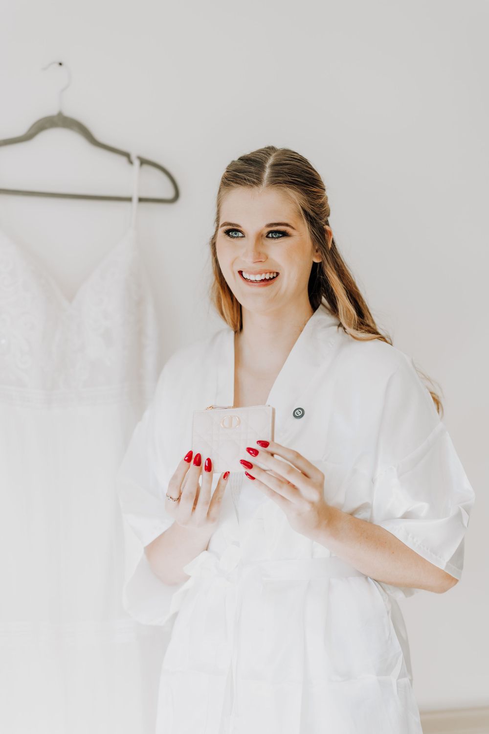 Bride in a white robe smiles while getting ready on their wedding day with red nail polish.