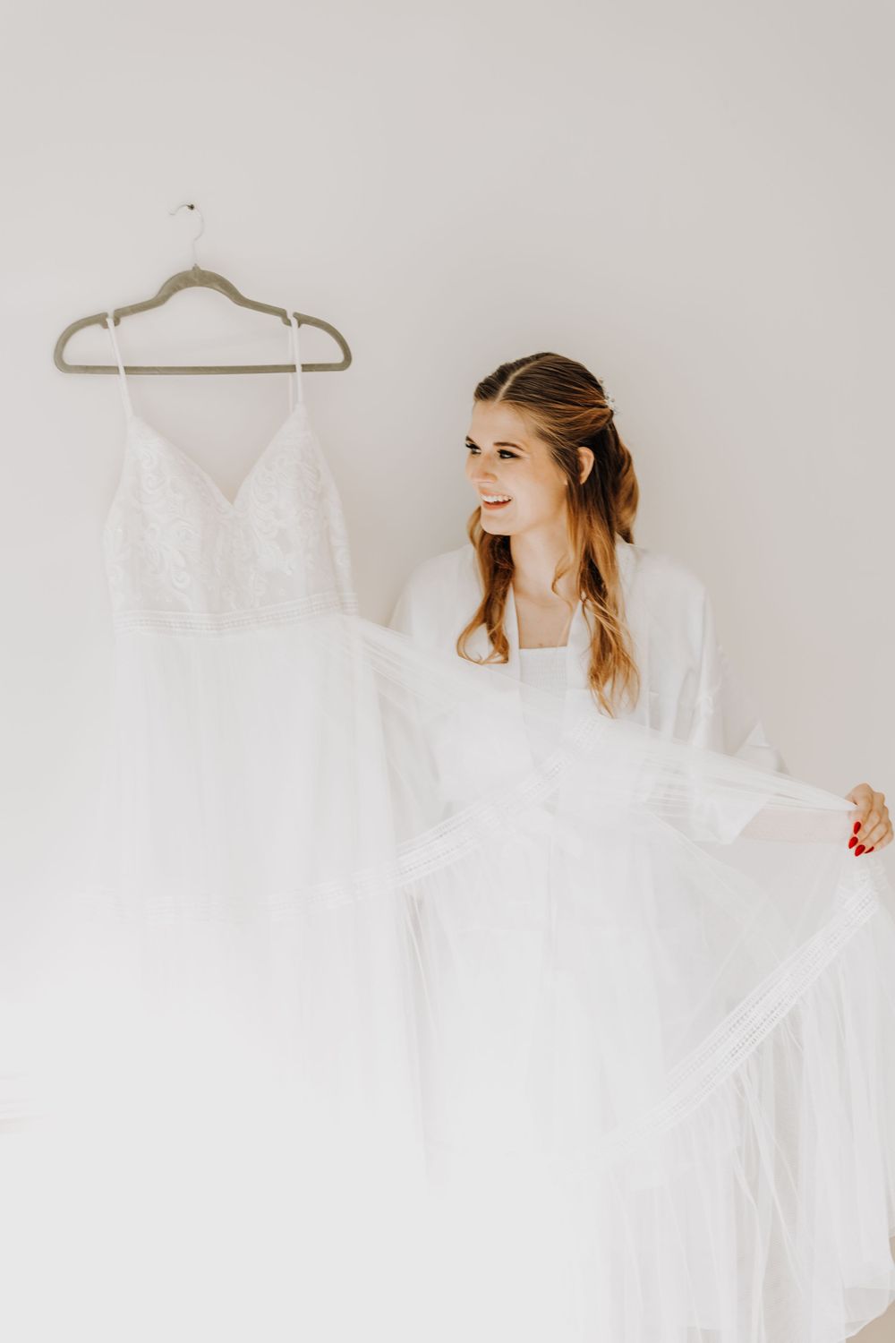 Bride in white robe admires a wedding dress hanging on display.
