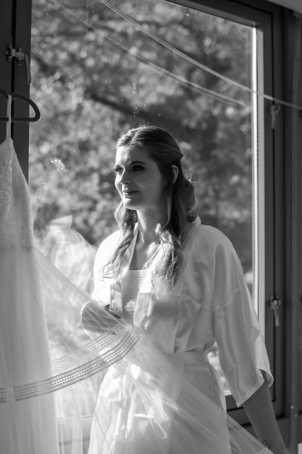 Bride stands by a window in a white robe during wedding preparations.
