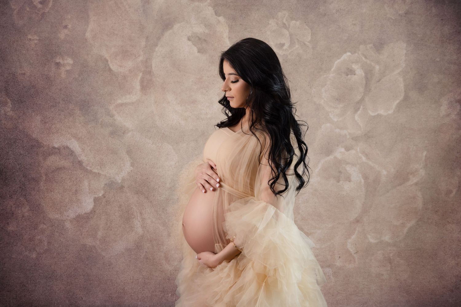 A pregnant figure in a cream-colored flowing dress poses gracefully against a textured mauve backdrop.