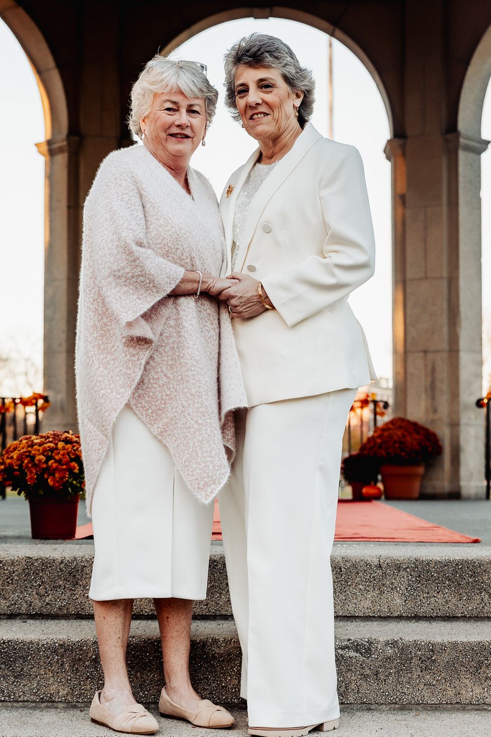 Two elegant women in white outfits embrace while standing in an archway decorated with autumn mums.