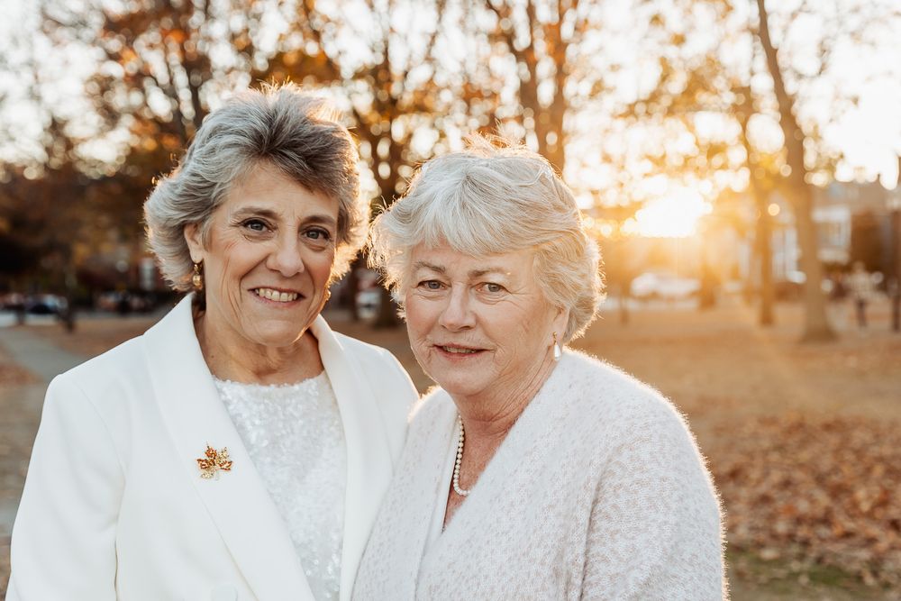A loving couple in crisp white outfits smile during golden hour with autumn leaves in the background.