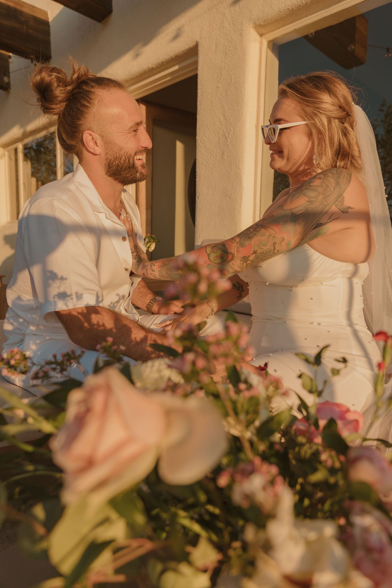 A couple laughing and sharing a romantic moment during sunset with pink flower decorations in the foreground.