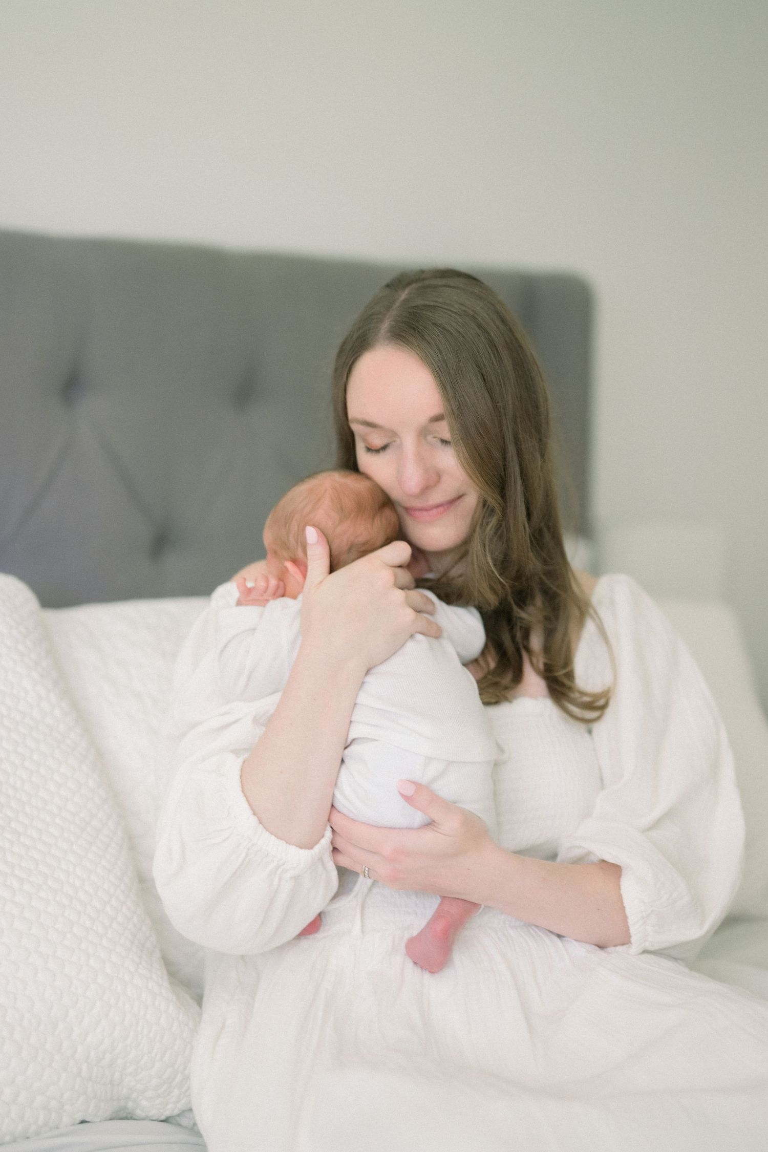 A tender moment captured in soft light as a mother in white cradles and gazes lovingly at her newborn baby on a gray bed.