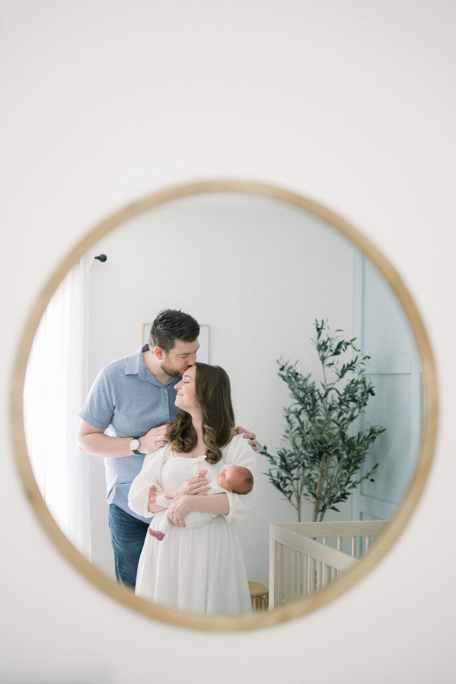 A tender family moment captured in a circular mirror reflection with a green plant in the corner of a bright nursery room.