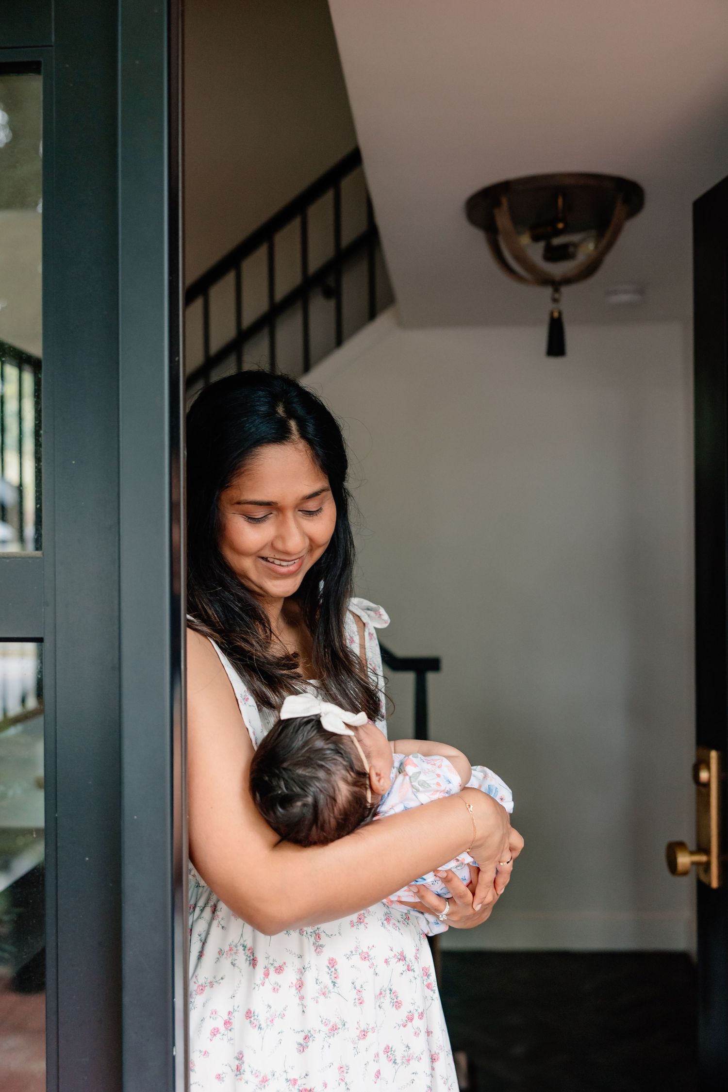 A woman in a floral dress stands in a doorway holding a sleeping baby wrapped in white fabric.