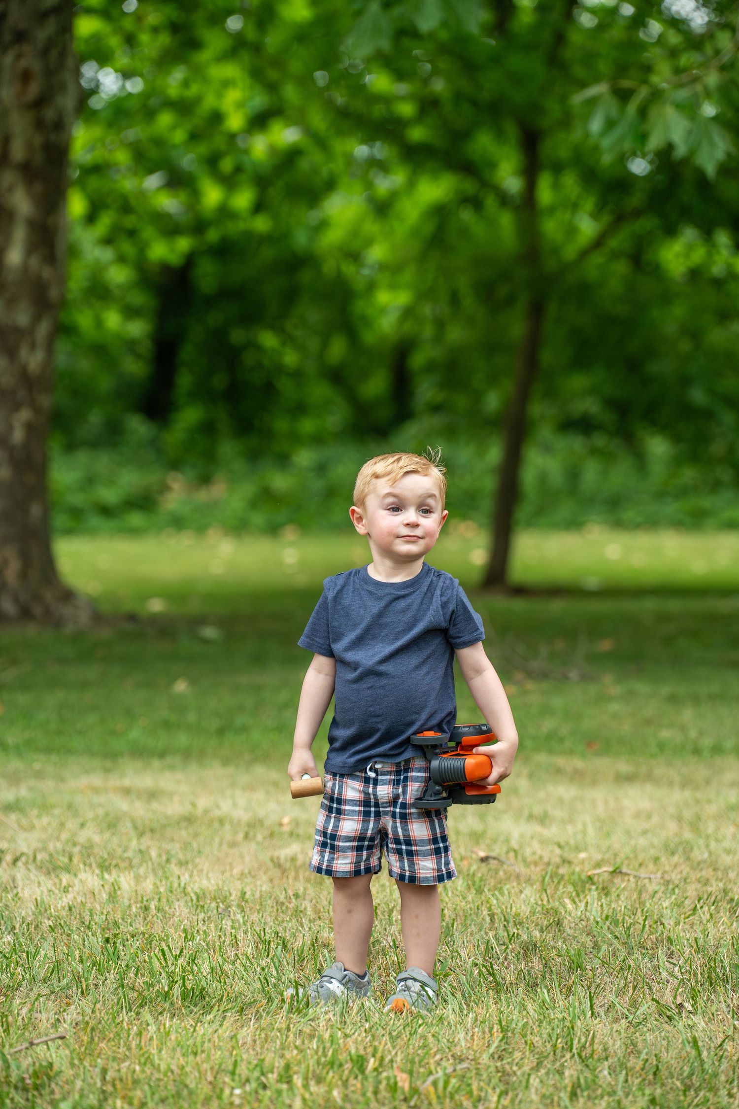 A child in plaid shorts plays on a grassy lawn surrounded by lush green trees on a sunny summer day. Boy has his favorite toy.