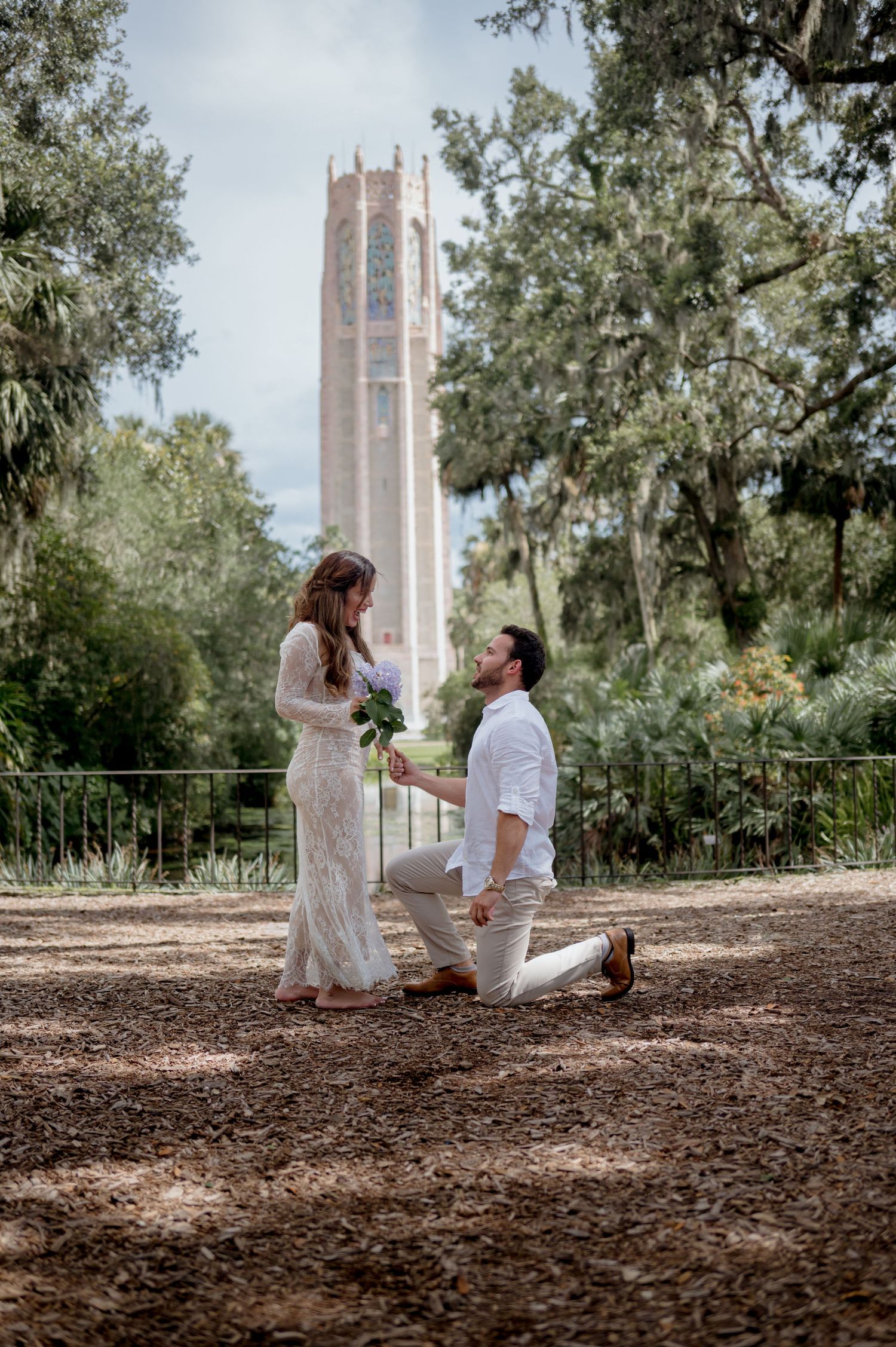 A proposal moment captured in sequence showing a couple in white clothing near a brick tower under shady trees.