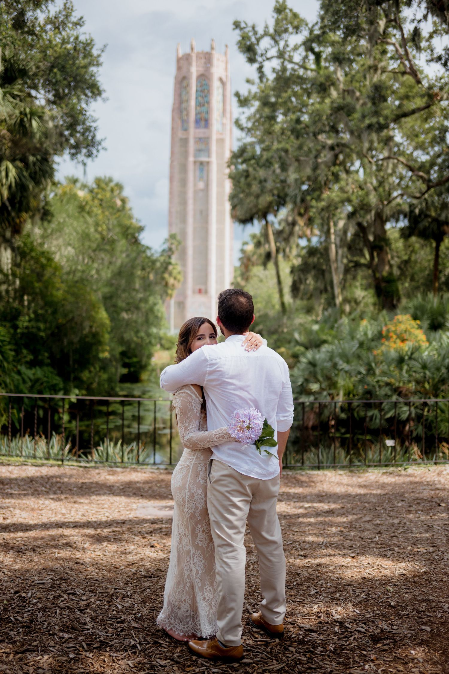 A couple embraces while looking at a tall bell tower surrounded by lush trees in a romantic outdoor setting.