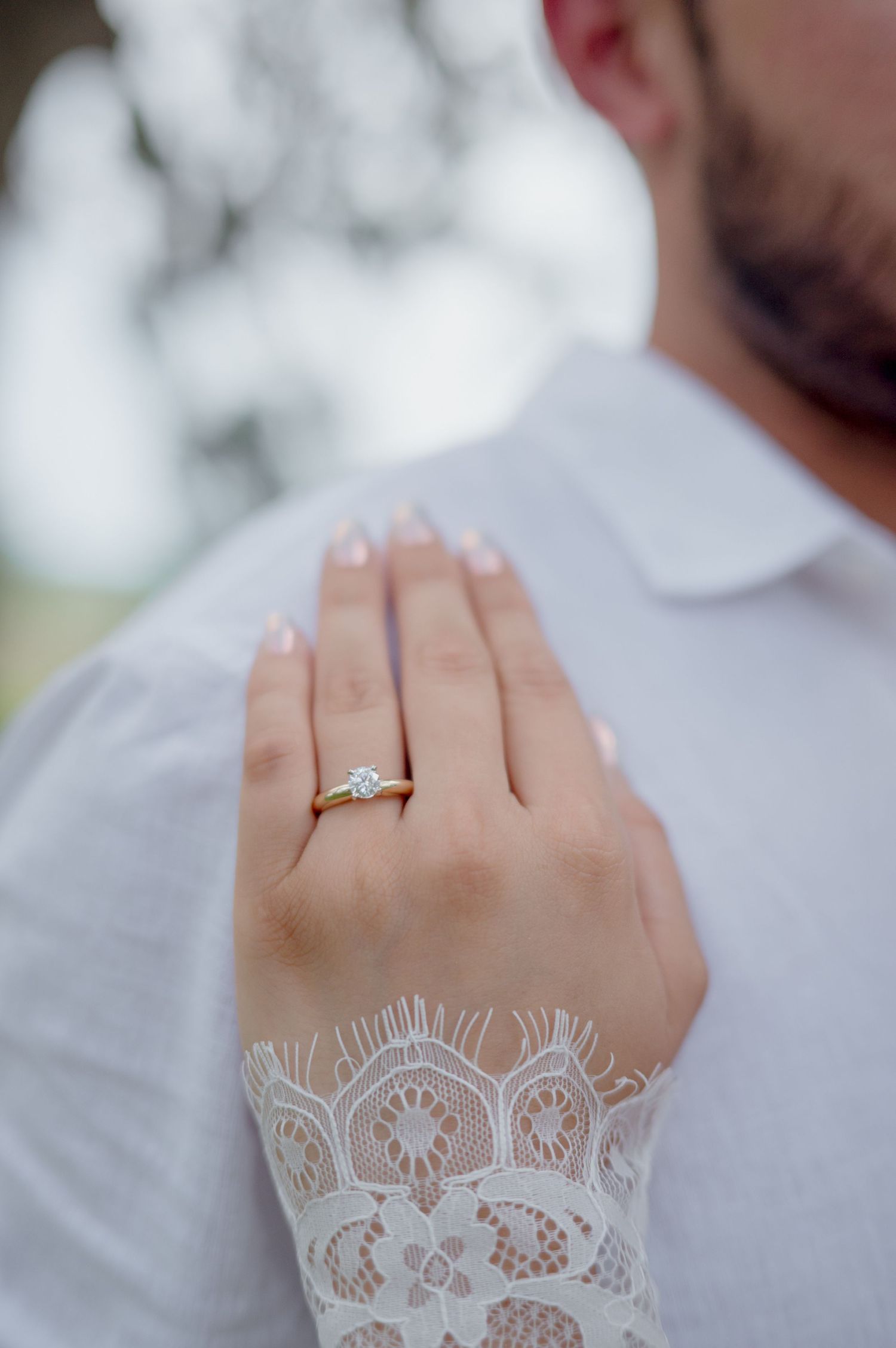 A delicate engagement ring sparkles on a hand wearing long lace sleeve detail against a soft blurred background.