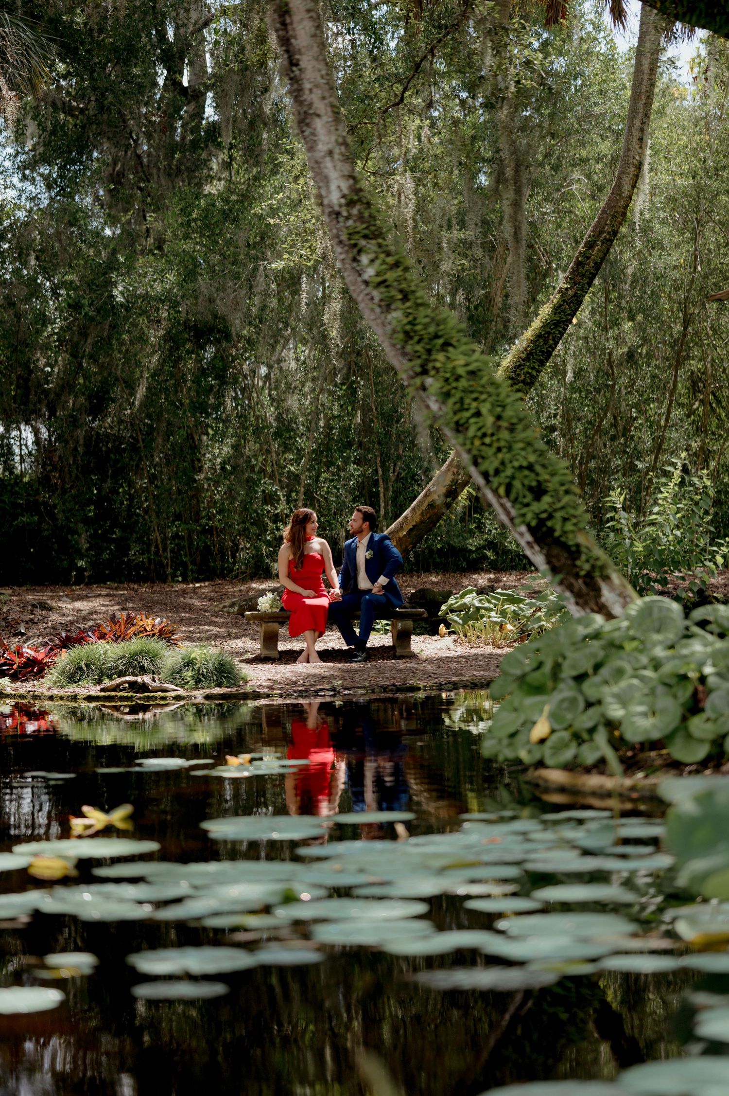 A moody forest lake scene with lily pads in the foreground and two figures in red and blue sitting by the water's edge.