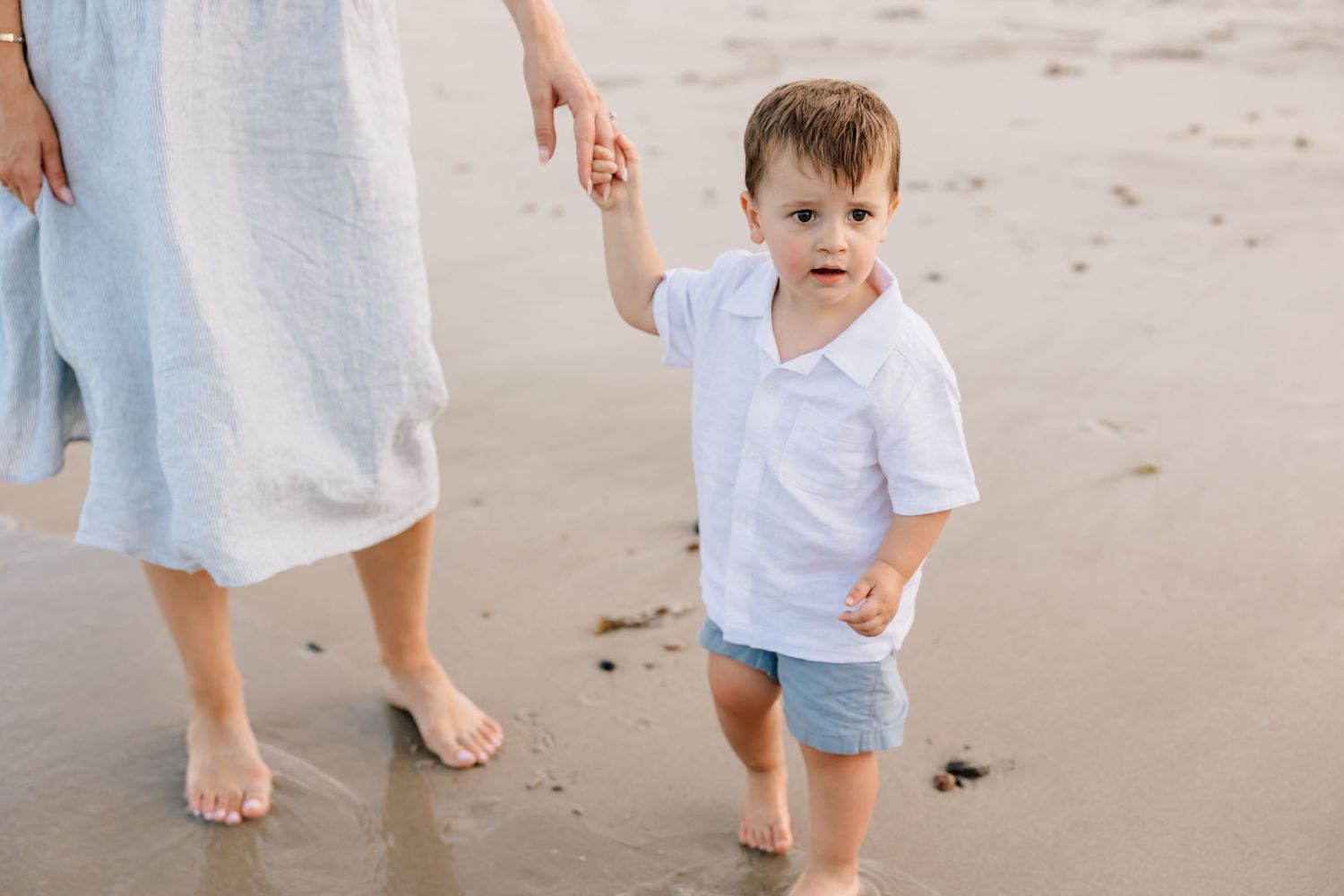 A small child wearing a white shirt and blue shorts holds hands with someone while walking on a sandy beach.