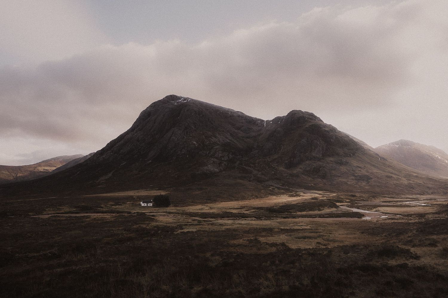 A moody mountain landscape in Glencoe the Scottish Highlands with dramatic clouds and a lone white building in the valley.