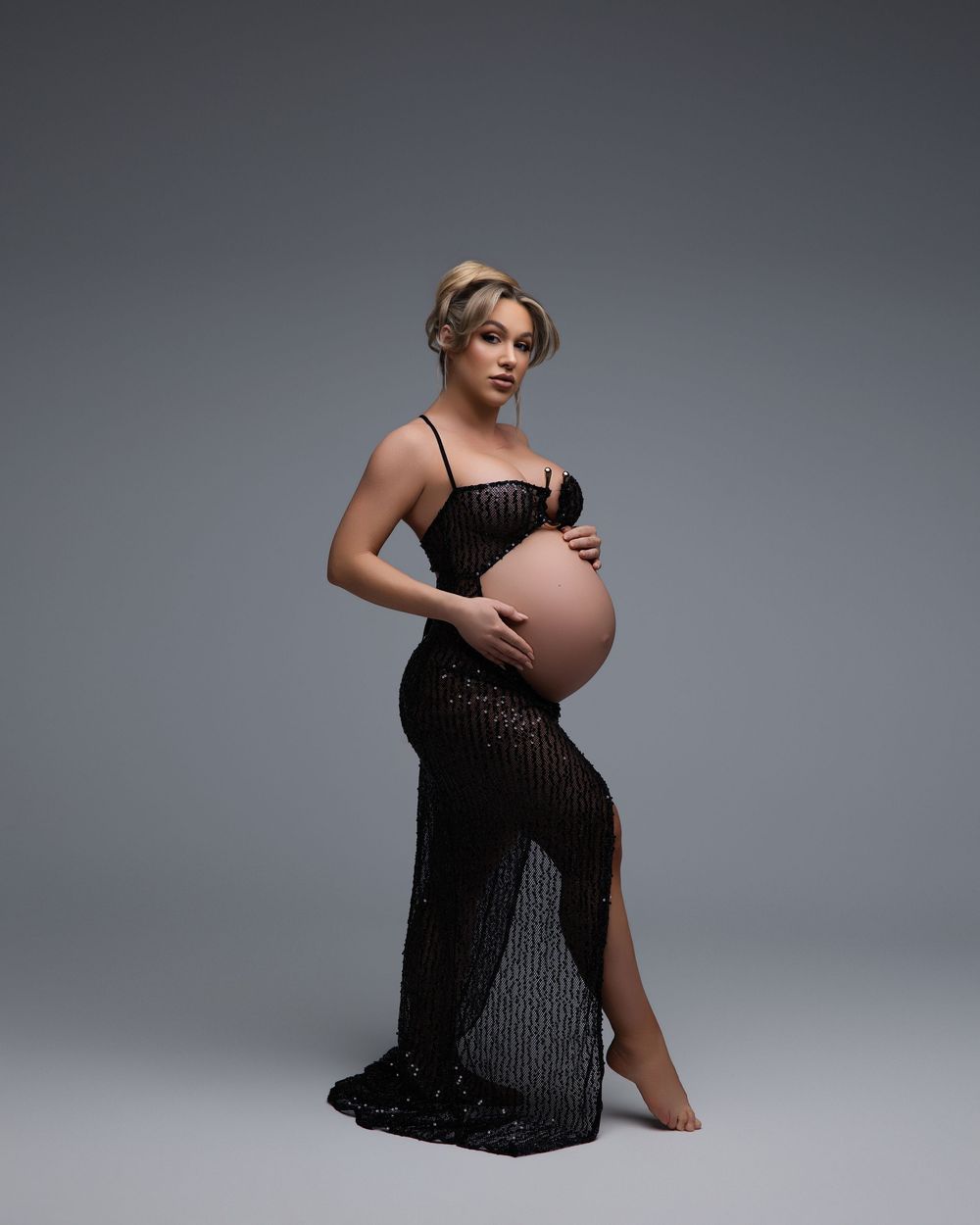 Person in a black sheer gown poses elegantly for a dramatic maternity photo against a gray studio backdrop.