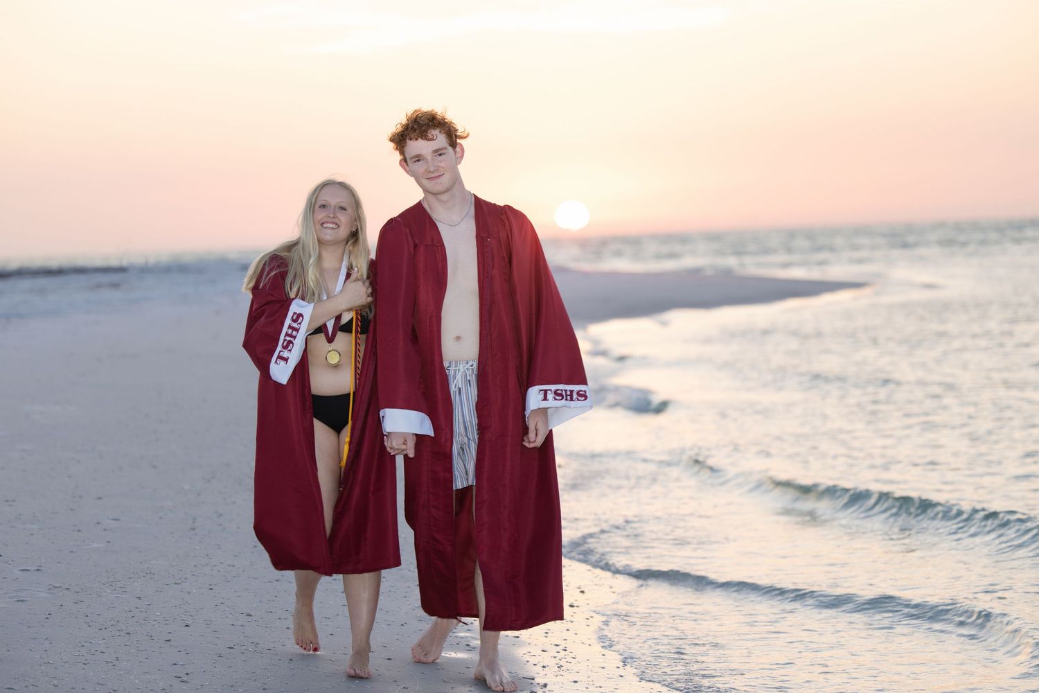 Two high school seniors in bathingsuits and graduation gowns walking on beach at sunset with waves breaking on shore in warm light.