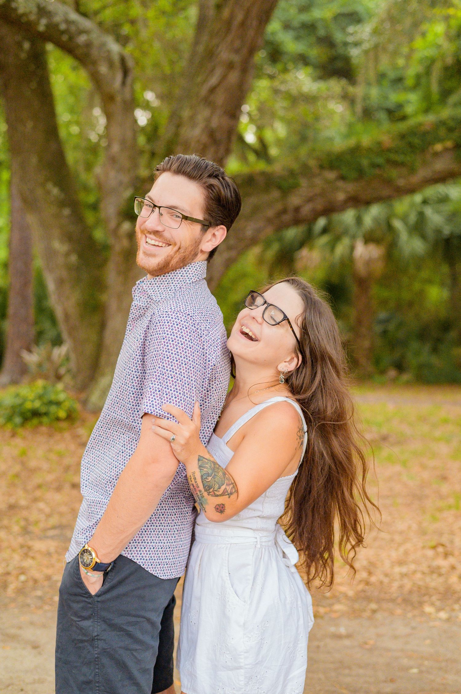 A playful outdoor portrait of a couple in glasses posing back-to-back in a park setting.