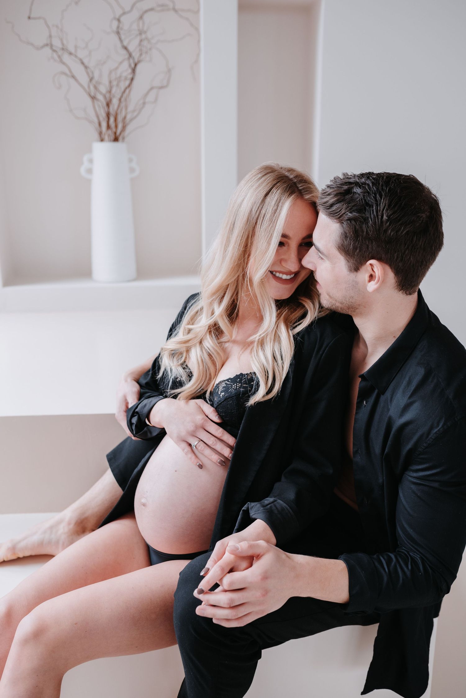 A romantic couple in matching black outfits shares an intimate moment while sitting together in a minimalist white room.