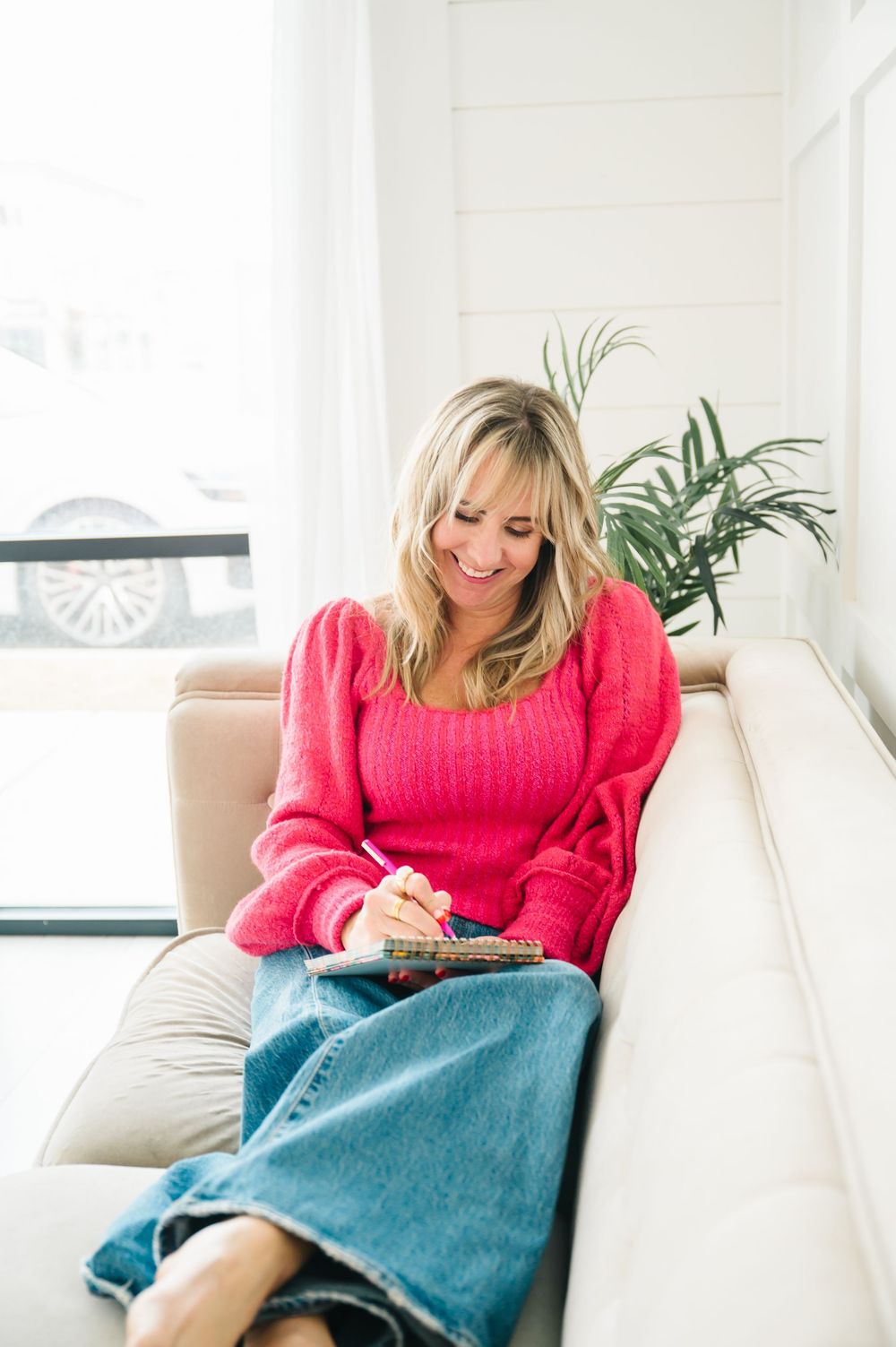 Someone wearing a bright pink sweater and denim relaxes on a white couch in a bright modern room.