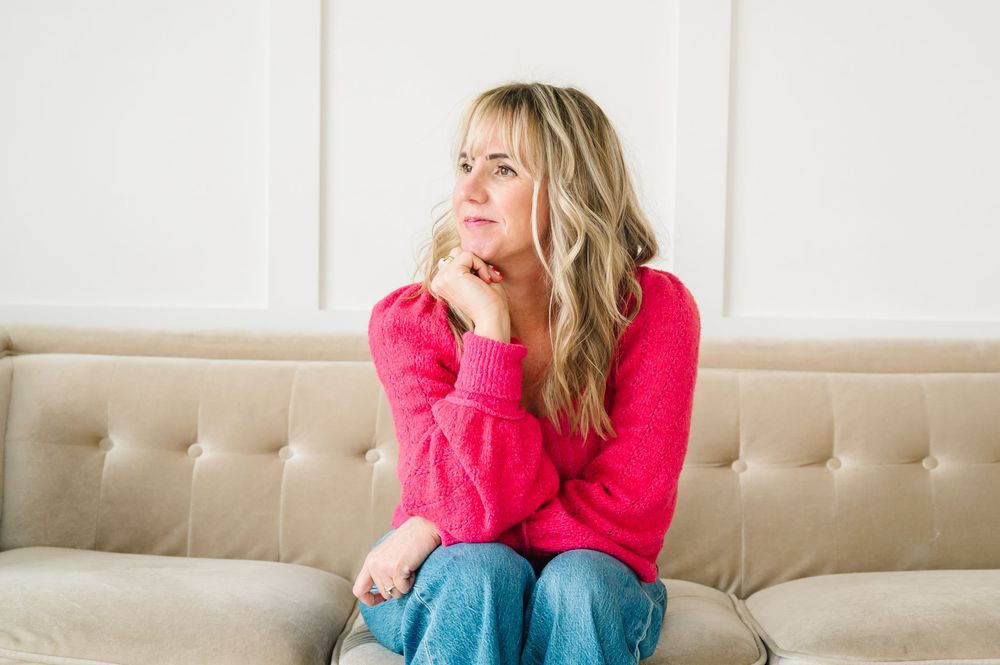 Woman in bright pink sweater sits on beige couch looking to the side while holding white coffee mug.