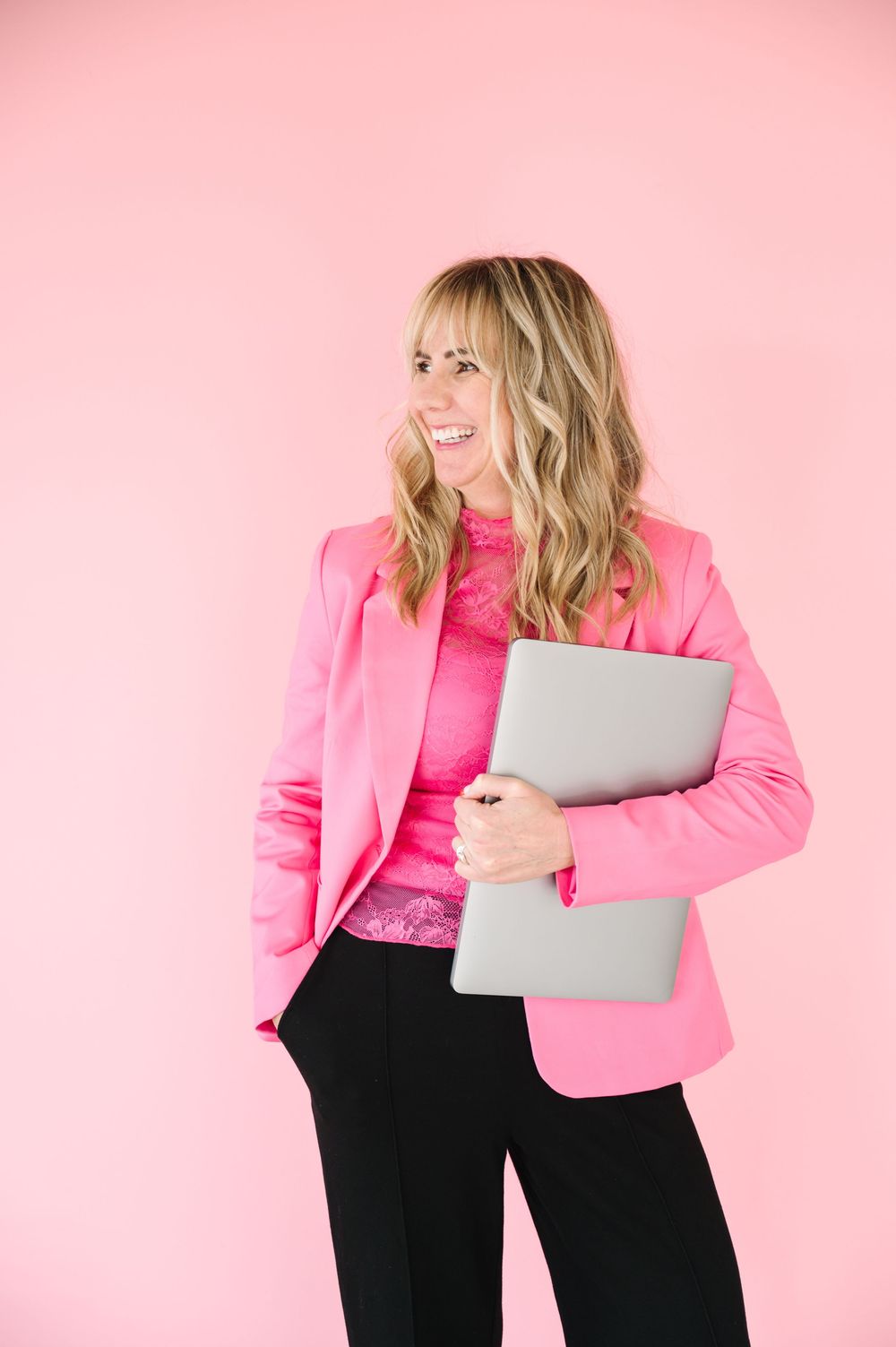 Professional wearing bright pink blazer jacket poses with laptop against pink background.