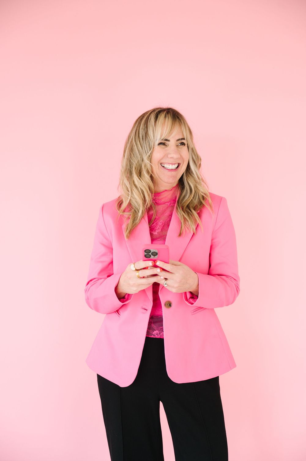 Woman in bright pink blazer and black outfit laughing and posing against pink background in professional photo series.
