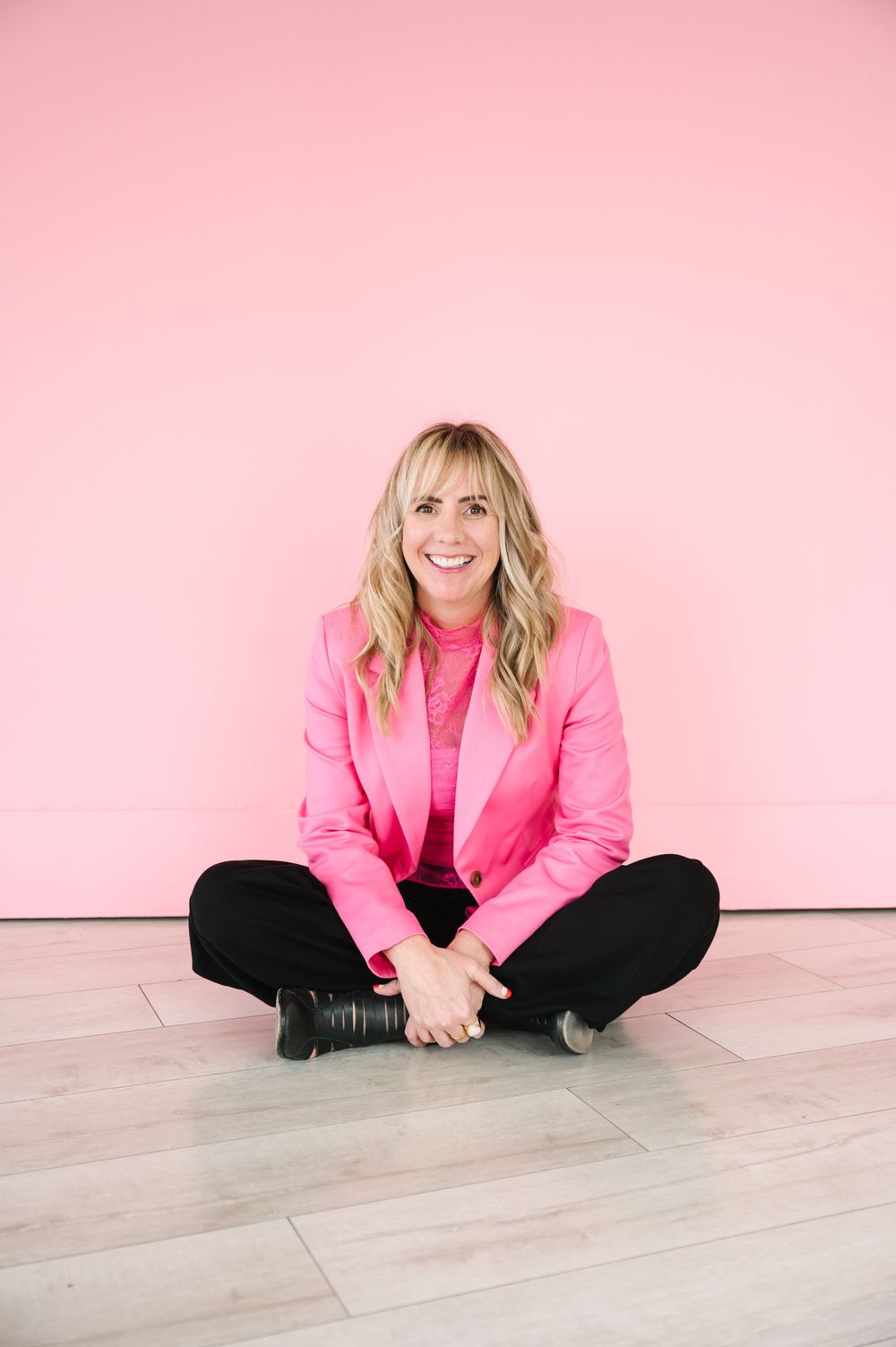 A person in a bright pink blazer and black pants sits cross-legged on a light wood floor against a pink wall.
