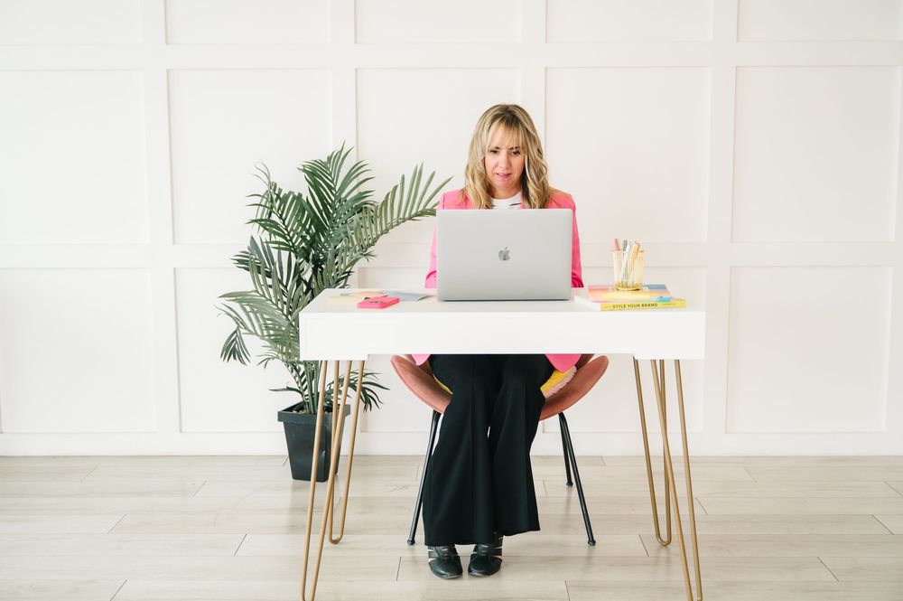 A person in business attire works at a minimalist desk with laptop, documents and indoor plants in a bright room.