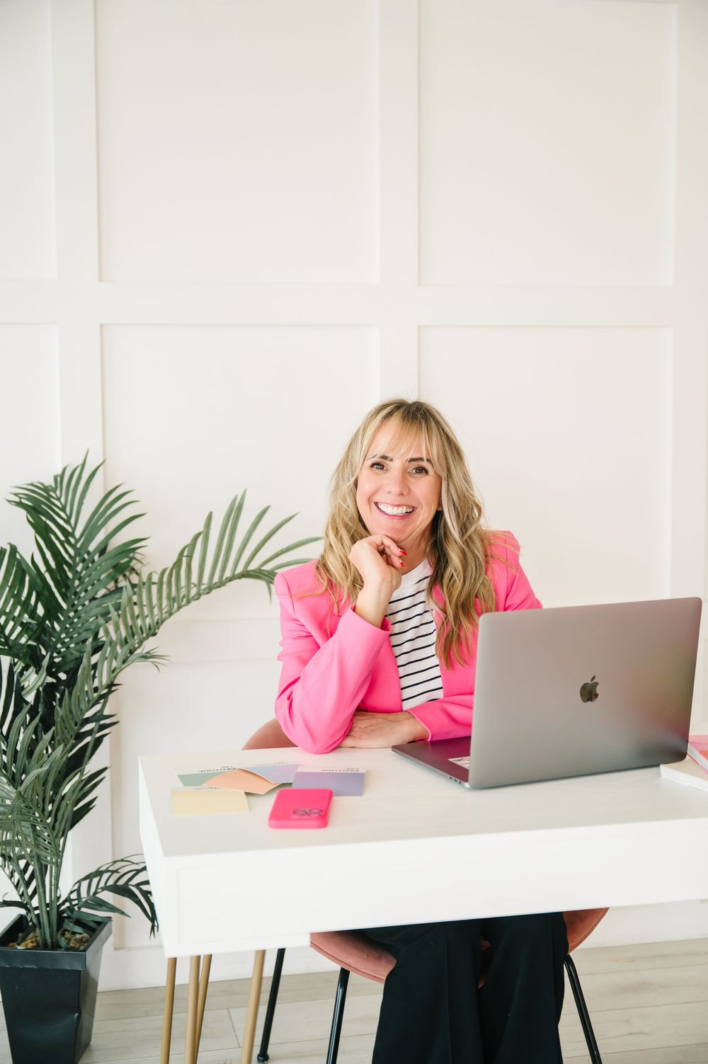 Person in bright pink blazer working at white desk with laptop and tropical plant decor in modern office setting.