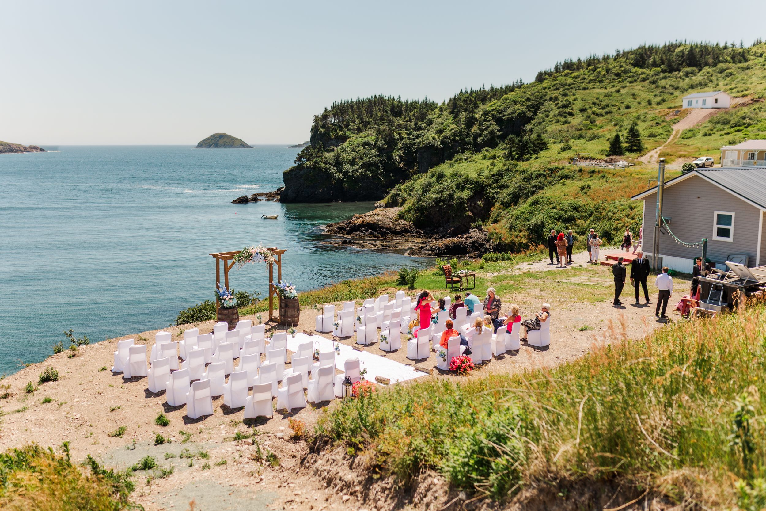 Outdoor beach wedding ceremony setup with white chairs and wooden arch overlooking the ocean and rocky coastline.