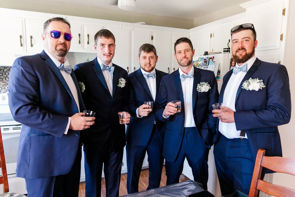 Wedding party members in navy suits pose together holding drinks for a celebratory photo.