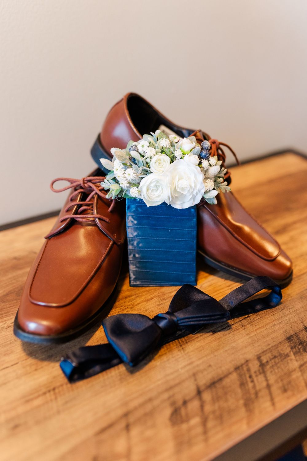 Brown leather dress shoes with a black bow tie and white floral arrangement in a navy blue vase on wooden surface.