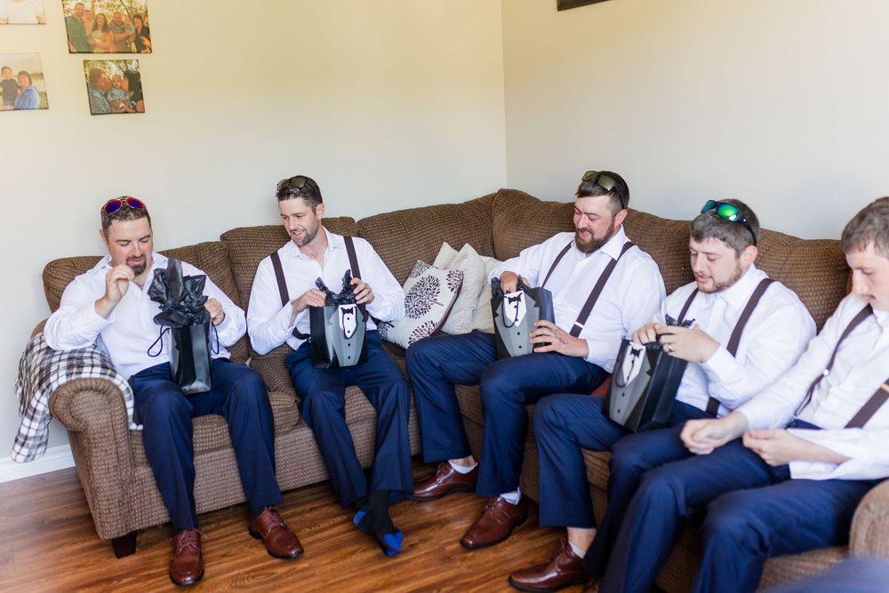 Groomsmen in navy blue suits and suspenders relax together on a couch while holding drinks.