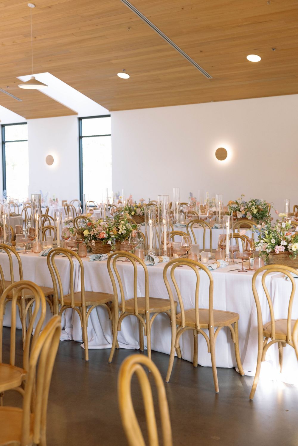Elegant wedding reception setup with wooden bentwood chairs, white tablecloths, and floral centerpieces in a modern venue.