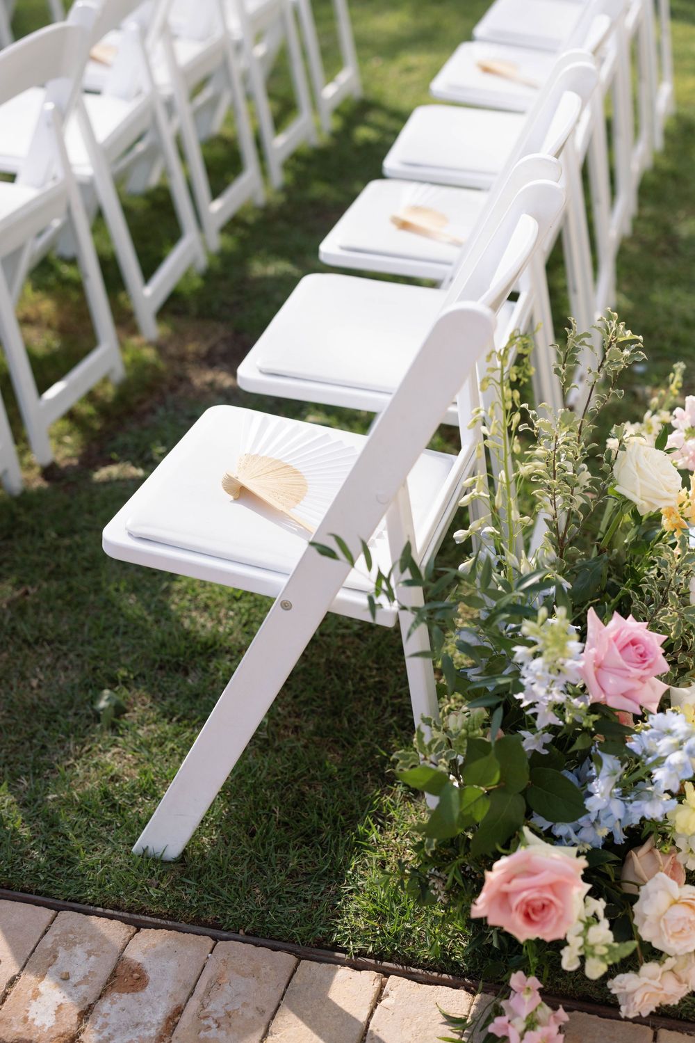 White wooden chairs adorned with pink and blue flowers line up for an elegant outdoor garden wedding ceremony.