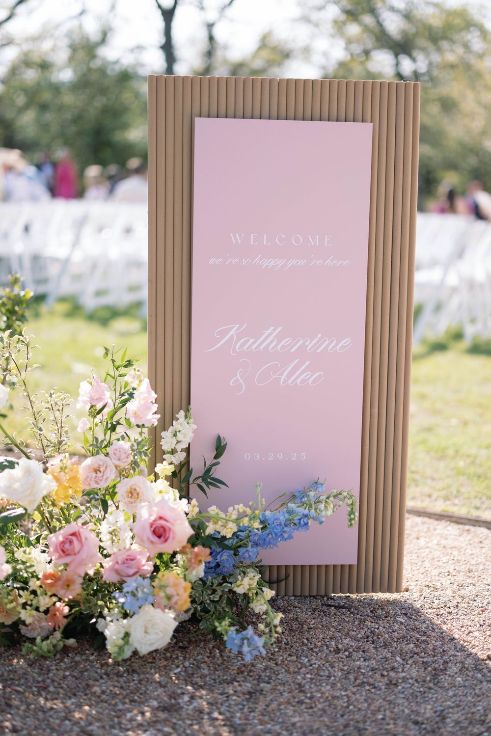 Elegant wedding welcome sign with pink lettering and floral arrangements featuring roses and blue delphinium flowers.
