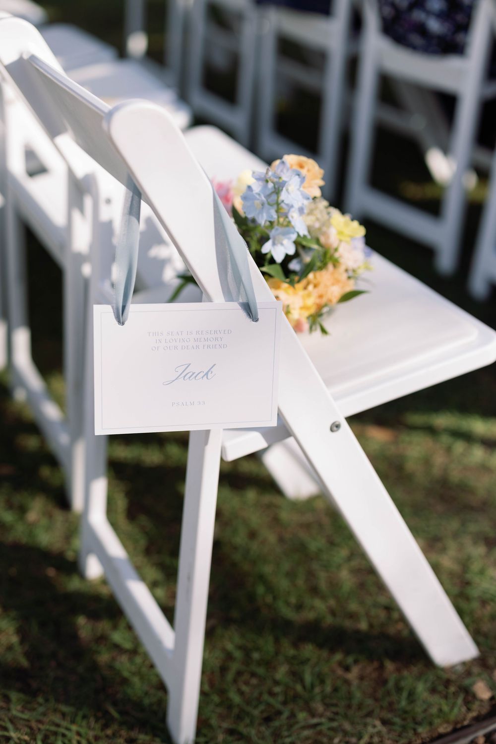 White folding chair with a small floral arrangement and invitation card at an outdoor wedding ceremony setup.