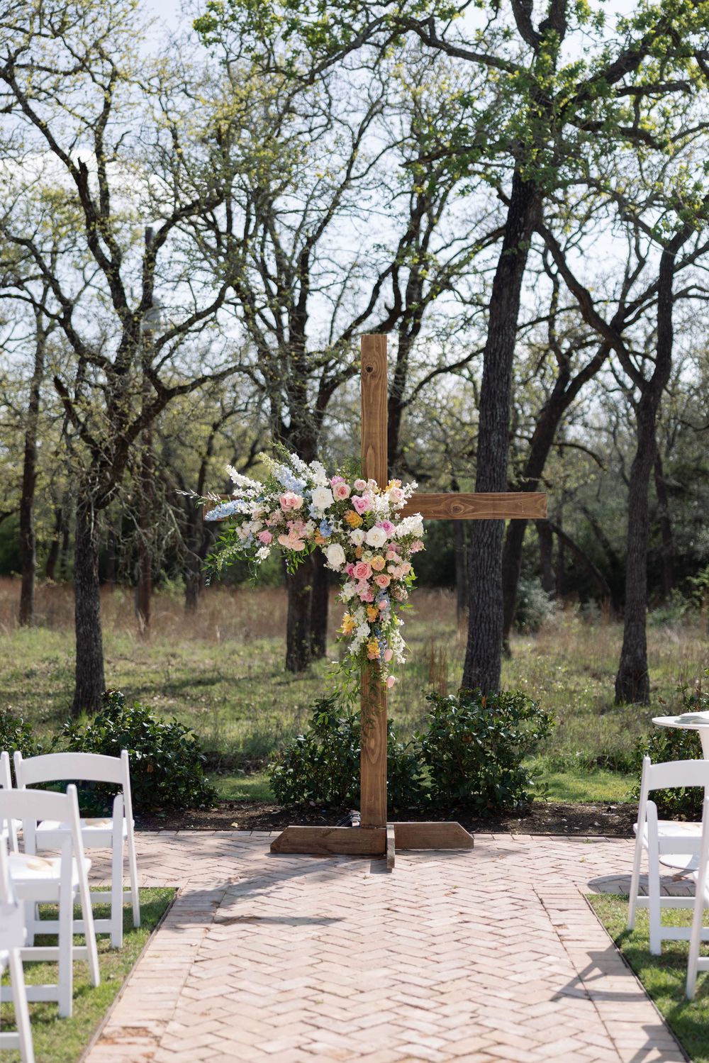 A wooden cross decorated with pink and white flowers stands at the end of a brick path with white chairs for an outdoor wedding ceremony.
